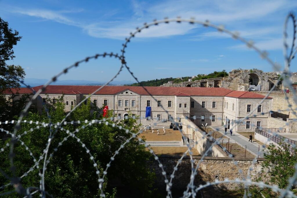 Avrupa Birliği desteğiyle yenilenen tarihi cezaevi avlusu, modern ziyaretçi yürüyüş yolları ve korunan orijinal tel örgülerin aslına uygun dokusu.